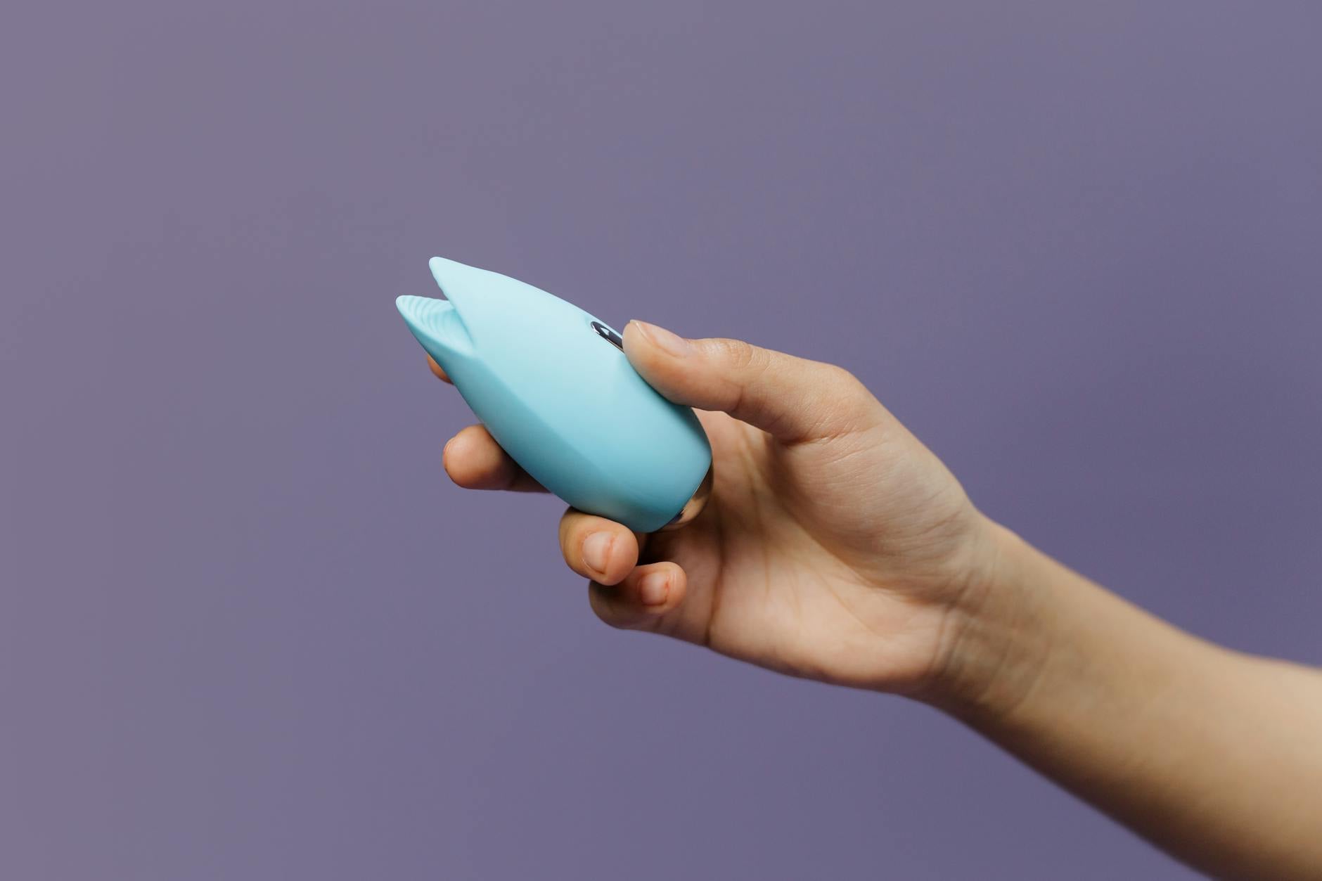A person holding two colorful clitoral vibrators against a purple background, showcasing modern pleasure tools