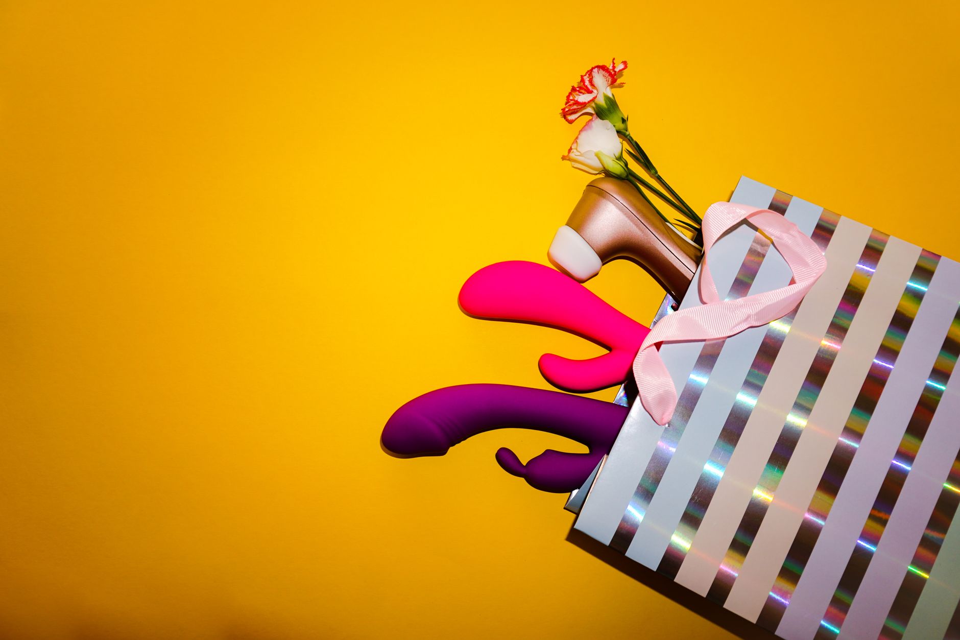 Colorful clitoral vibrators and flowers in a holographic gift bag on a bright yellow background